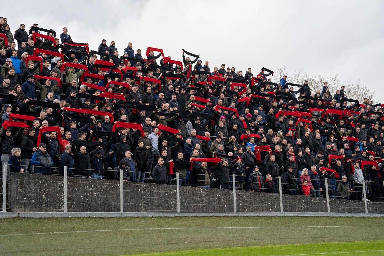Une tribune pleine de supporters, écharpes rouges et noires levées, vue depuis le bord de la pelouse en plein jour